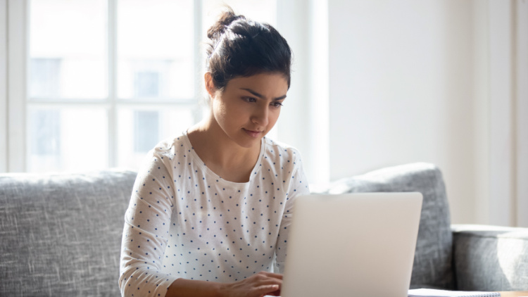 A woman looks at her laptop computer with a focused expression, sitting at a desk in front of a window