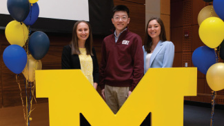 Three students stand on a stage behind a large block M prop with maize and blue balloons beside them