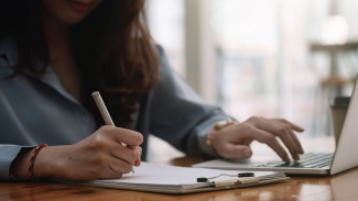 A woman types on her computer at a table while writing notes on a paper on a clipboard with a pen